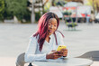 © Nana_studio - a smiling young African American girl with pink hair sitting at a table in an outdoor cafe and using a mobile phone on a sunny day.