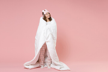 Wall Mural - Full length portrait of smiling pretty cute young woman in pajamas home wear sleep mask wrapping in blanket looking aside resting at home isolated on pink background studio . Relax good mood concept.