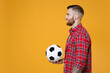 Side view smiling young man football fan in basic red shirt cheer up support favorite team with soccer ball looking aside isolated on yellow background studio portrait. People sport leisure concept.