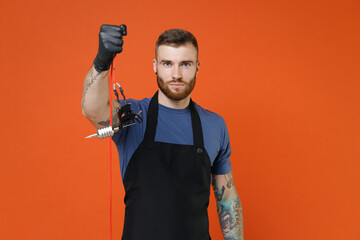 Young professional tattooer master artist tattooed man in blue t-shirt apron hold machine black ink in jar, equipment for making tattoo art on body isolated on brown background studio portrait.