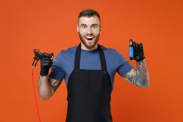 Surprised young professional tattooer master artist tattooed man in t-shirt apron hold machine black blue ink jar equipment for making tattoo art on body isolated on brown background studio portrait.