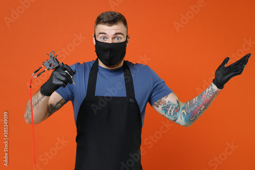 Shocked professional tattooer master artist tattooed man in t-shirt apron face mask hold machine black ink in jar equipment for making tattoo art on body spreading hands isolated on brown background.