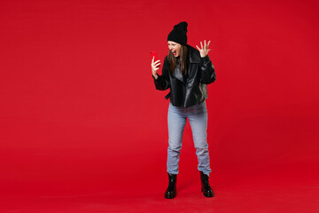 Wall Mural - Full length of angry irritated young brunette woman 20s in black leather jacket white t-shirt hat talking on mobile phone screaming swearing spreading hands isolated on red background studio portrait.