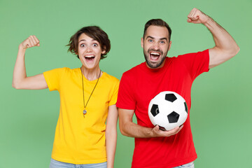 Canvas Print - Excited strong couple friends sport family woman man football fans in yellow red t-shirts cheer up support favorite team with soccer ball showing biceps muscles isolated on green background studio.