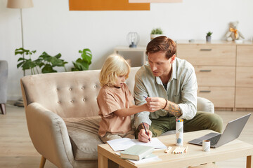 Father helping to his little son doing homework they sitting on sofa at the table in the room