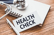 © Nastassia - Health check - text on business card on wooden table with stethoscope and card for medical records.