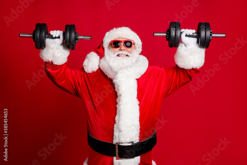 Photo of pensioner old man white beard lift heavy dumbbells smiling leave youngsters behind close strength competition win wear santa costume sunglass headwear isolated red color background