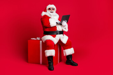 Full length photo of retired old man white beard sit giftbox hold netbook search way north pole wear x-mas santa costume glove coat belt sunglass cap boot isolated red color background