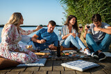Fototapeta  - Group of friends having fun on picnic near a lake, sitting on pier eating and drinking wine.