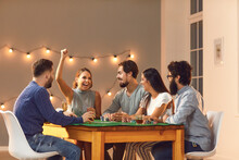Excited Young Woman Celebrating Poker Victory Sitting At Table With Friends At Casino Themed Party