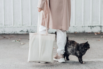 Wall Mural - Woman in coat with white cotton bag in her hands and cute cat sitting near. Mockup and zero waste concept.