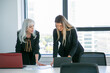 © Mangostar - Serious businesswomen discussing project in meeting room, standing at table, watching content on laptop. Front view. Business communication concept