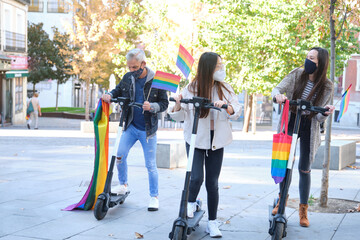 Three young caucasian and asian homosexual friends riding scooters with LGBT flags wearing face masks. LGBT pride celebration in pandemic times.