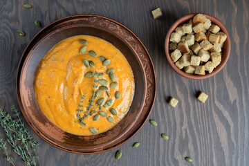 Wall Mural - Homemade vegetarian pumpkin soup of orange color made of pureed vegetables cooked on thanksgiving decorated with seeds and thyme served in bowl on dark wooden background with croutons. Horizontal
