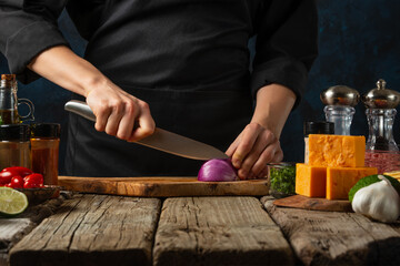 Wall Mural - Professional chef in black uniform cuts with knife onion on chopped board for cooking filling for tacos. Backstage of preparing traditional mexican recipe. Concept of tasty food. Dark background.