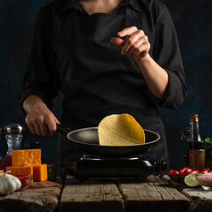 Wall Mural - Professional chef in black uniform frying corn tortillas in pan on dark blue background. Backstage of preparing traditional mexican tacos at wooden rustic table. Concept of tasty street food.