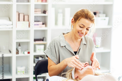 Beautiful happy caucasian woman smiling while doing face massage to client at spa - Female beautician doctor cosmetologist applying face mask at salon - healthcare concept copy space