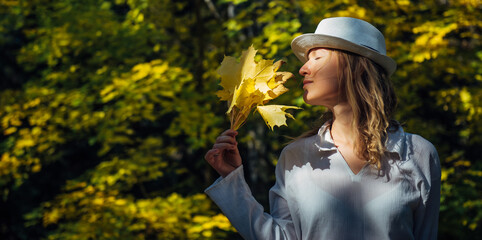 Wall Mural - Elegant young blonde in white shirt and hat in autumn park on sunny day. Beautiful happy smiling woman held a bouquet of yellow maple leaves to her face. People in outdoor leisure activity.