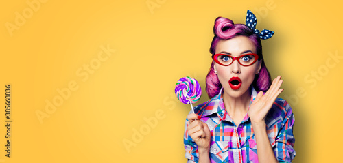Purple head excited very surprised woman with lollipop. Pinup girl in red glasses, with wide opened mouth, eyes. Beauty model at retro and vintage concept. Yellow orange background with copy space. © vgstudio