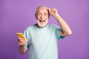 Photo portrait of elderly man lifting up glasses holding phone in one hand isolated on vivid purple colored background