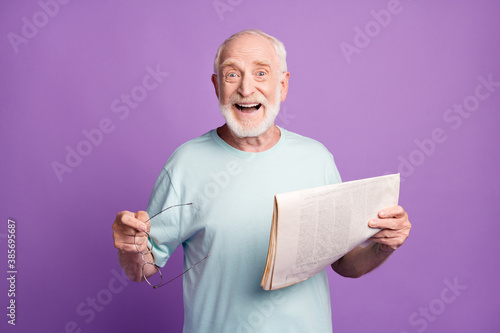 Portrait of happy smiling grey hair beard pensioner hold glasses newspaper wear light blue t-shirt isolated on violate background