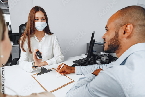 Fototapeta Attractive woman car seller wearing mask gives keys to car buyer