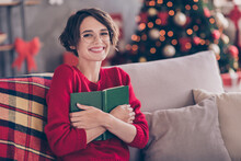 Photo Of Pretty Young Lady Cuddle Paper Book Toothy Smile Sitting Sofa Look Camera Wear Spectacles Red Pullover Indoors