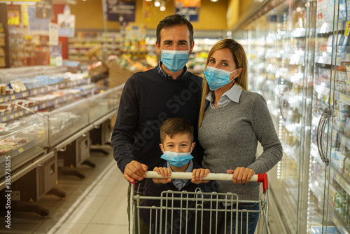 Authentic shot of happy family of father, mother and son wearing medical masks to protect themselves from disease are looking in camera while shopping for groceries together in supermarket.