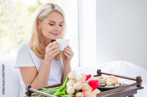 Photo of charming aged lady sit bed white linen drink smell fresh coffee beverage dreamer bedroom indoors