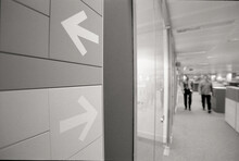 Close-up Of Arrow Symbol On Wall Against Colleagues Walking In Corridor At Office