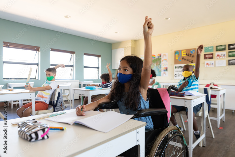 Disable girl wearing face mask raising her hand while sitting on wheelchair in class - obrazy, fototapety, plakaty 