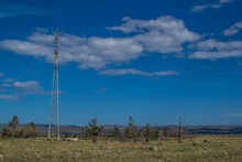 Power Pole Stands Among Coniferous Green Pine Trees On Dry Yellow Grassy Steppe Of Lake Baikal Nature. Blue Mountains Background. Blue Sky. Summer Landscape