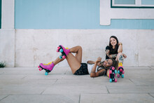 Smiling Female Friends With Roller Skates Relaxing On Footpath Against Wall In City