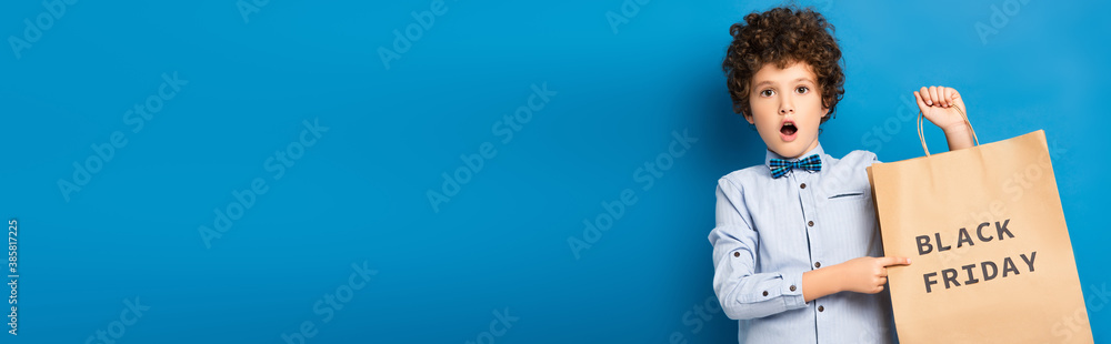horizontal image of shocked and curly boy pointing with finger at shopping bag with black friday lettering on blue - obrazy, fototapety, plakaty 
