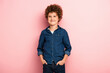 pleased and curly boy in denim shirt standing with hands in pockets on pink