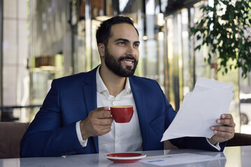 Wall Mural - Portrait of young handsome arabic manager working with documents, sitting at workplace. Smiling middle eastern businesswoman reading contract, planning start up, holding cup of coffee in modern office