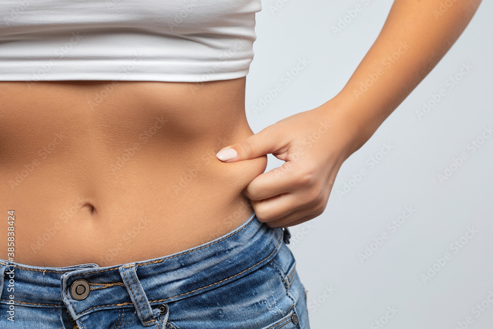 Girl pulls the skin on the abdomen, showing the body fat in the abdominal area and sides. Treatment and disposal of excess weight, the deposition of subcutaneous fat. - obrazy, fototapety, plakaty 