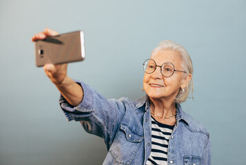 Wall Mural - Happy smart old lady in blue jeans jacket, black and white striped shirt smiles and takes selfie holding mobile phone at arm length. Online communication for elderly people concept.