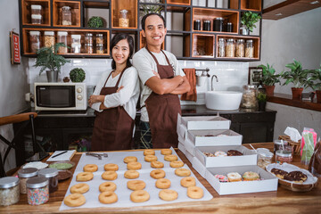 Confident Asian couple standing in a kitchen with crossed hands in front of donuts on the table