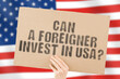 © AndriiKoval - The question ' Can a foreigner invest in USA? ' on a banner in men's hand with blurred American flag on the background. Investment. Business. Money. Rules. Policy. Legislation. Law
