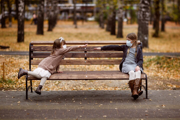 Children two girls friends for a walk in the park, children sit on a bench wearing masks, social distance and friendship during quarantine and restrictions