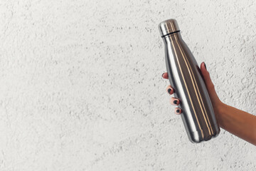 Close-up of female hand holding reusable steel thermo water bottle on background of textured wall.