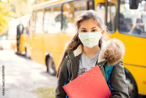 Secondary school girl learner in protective mask in front of school bus, ready to go home by bus. School education during the covid-19 pandemic