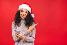 Happy Excited African American Woman In Red Santa Claus Hat With Mobile Phone Isolated Over Red Background. Pointing Finger.