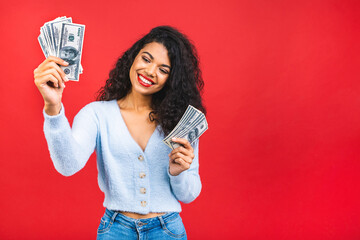 Portrait of a cheerful young curly african american woman holding money banknotes and celebrating isolated over red background.