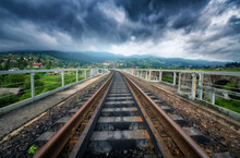 Railroad Bridge In Mountains In Overcast Day In Summer. Railway Station In Village At Moody Sunset. Industrial Landscape With Railway Platform, Green Trees And Grass, Dramatic Cloudy Sky, Buildings