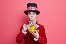 Surprised Man With Colorful Makeup Dressed In Mad Hatters Costume Enjoys Drinking Tea On Party Dresses For Halloween Poses Against Pink Background. Costumed Carnival Or Parade. Wonderland Character