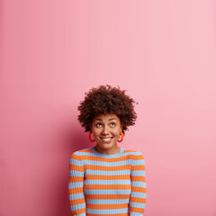 Vertical shot of happy curious woman with Afro hair bites lips and looks upwards dressed in casual jumper isolated over pink background. Glad female has satisfied expression notices something nice