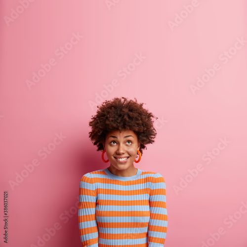 Vertical shot of happy curious woman with Afro hair bites lips and looks upwards dressed in casual jumper isolated over pink background. Glad female has satisfied expression notices something nice