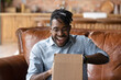 © fizkes - Overjoyed young African American man unpack cardboard box triumph with good quality order. Happy 20s biracial male unbox carton package with internet delivery, shopping on web online from home.